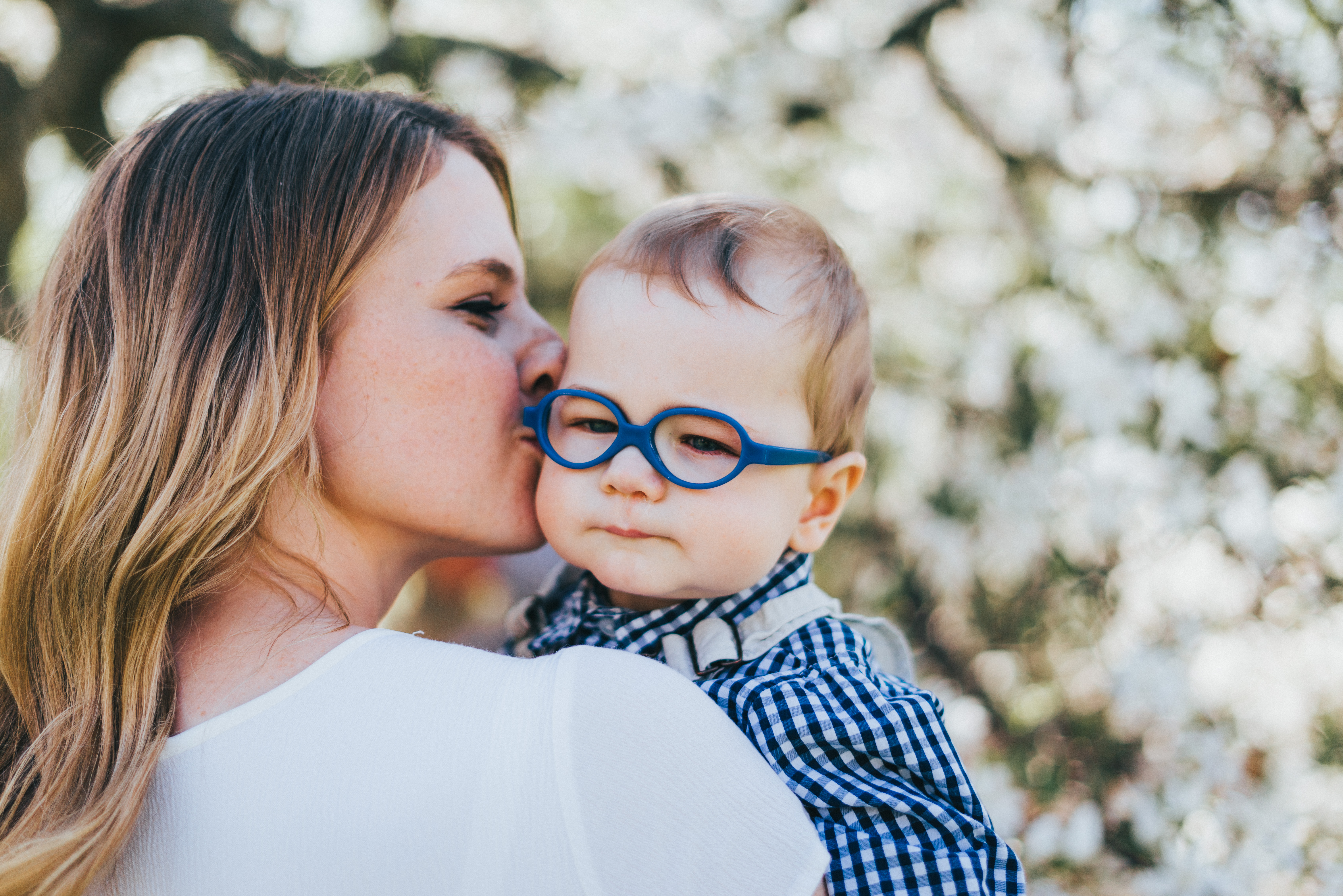 Author kissing her infant son with vision impairment on the cheek. She is in a white dress with curled hair and he is wearing dark blue baby glasses in a matching blue gingham dress shirt.
